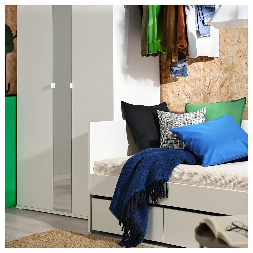 White daybed with blue blanket, black and green pillows against grey wall with clothes.