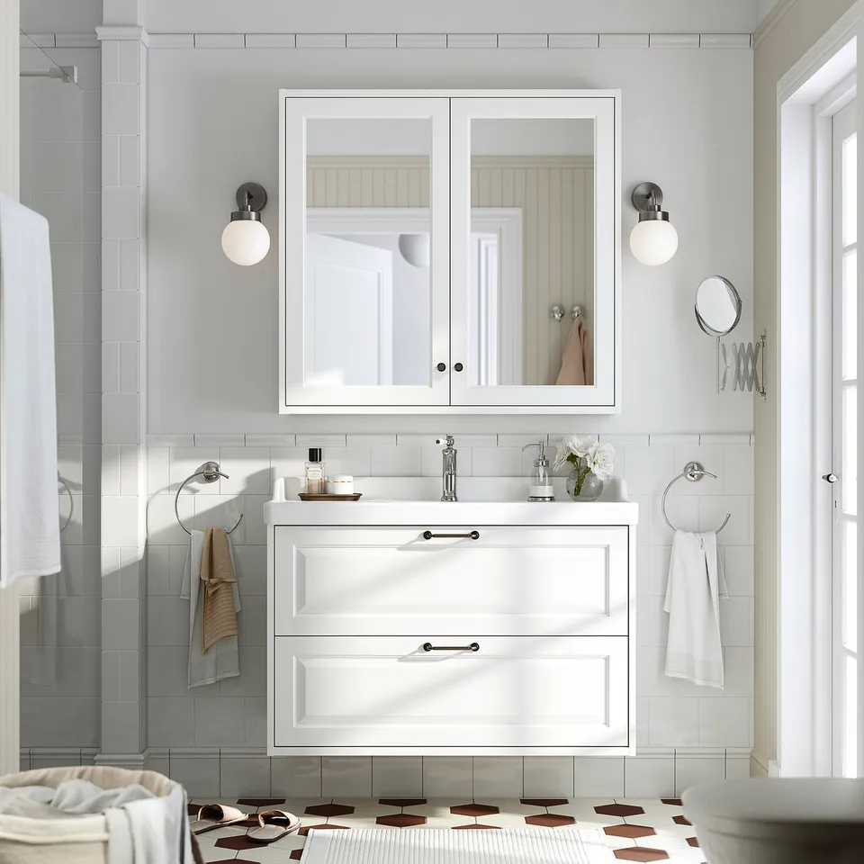 TÄNNFORSEN bathroom cabinet with mirrored doors, lights, and towels. Vase with flowers on vanity. Minimalist, bright and elegant.