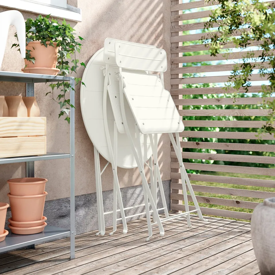 Three white SUNDSÖ folding chairs stacked neatly on a wooden balcony. Regional plants and decorative pots nearby.