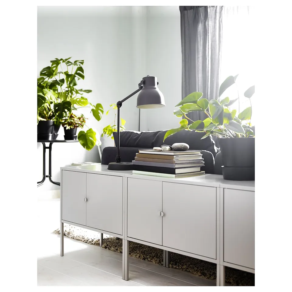 White side cabinet with grey handles, lamp, books, plants.