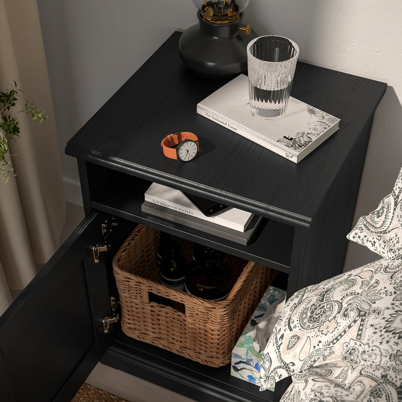 Black bedside table with open compartment, containing a woven basket and bottles. Top holds books, watch, and glass.