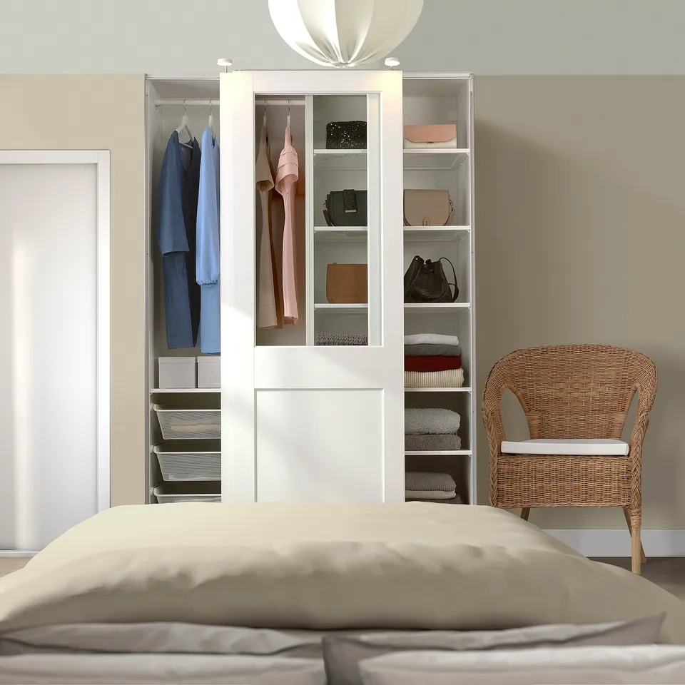 White PAX wardrobe with glass doors, beside bed and chair, holding clothes and bags.