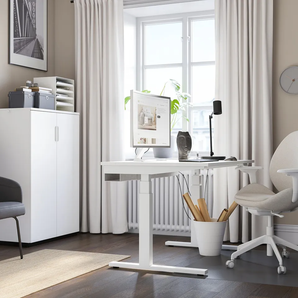 Modern minimalist home office: white MITTZON sit-stand desk with monitor, lamp, plant. Comfortable chair, storage, rug, large window.