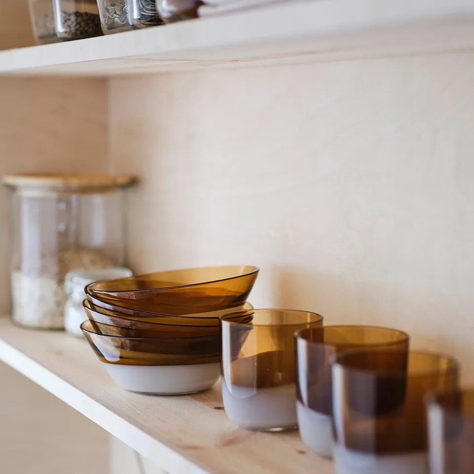 Shelf with stacked amber-colored glass OMMJÄNGE bowls, smaller than clear jars with wooden lids.