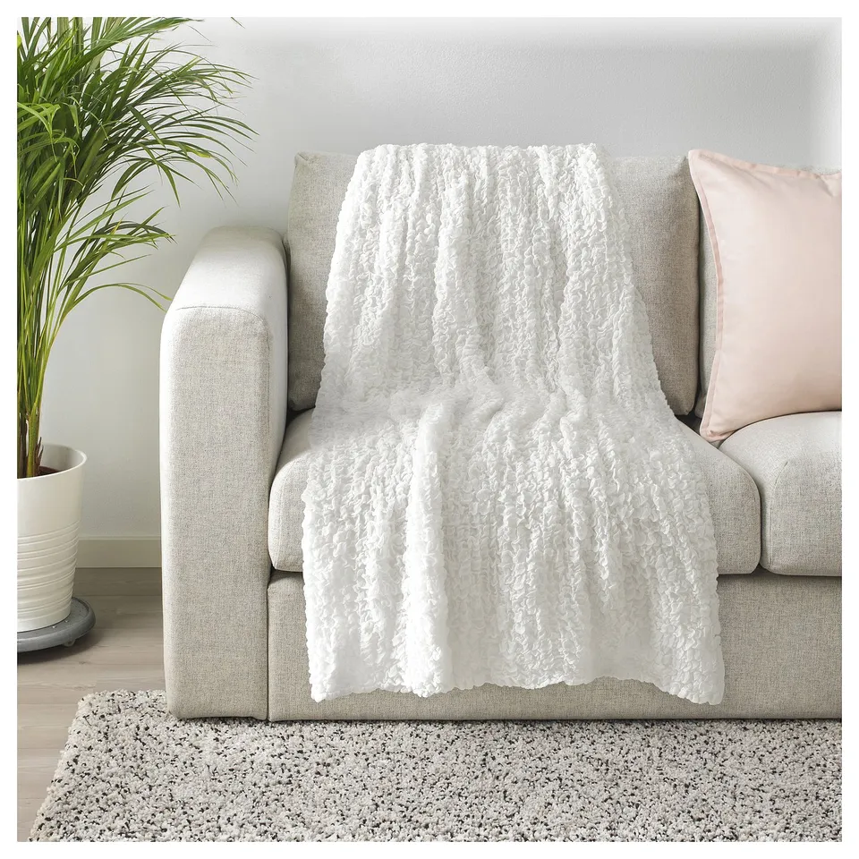 A white, textured OFELIA throw blanket draped over a beige sofa. The blanket is soft and made from 100% recycled polyester.