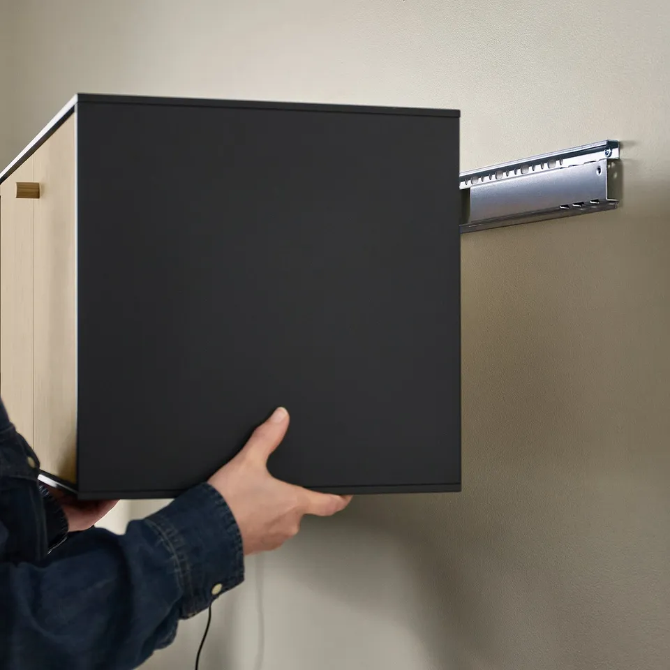 Person installing a black BOASTAD wall shelf, featuring thin lines and oak veneer, with wall-mounted metal rail visible.