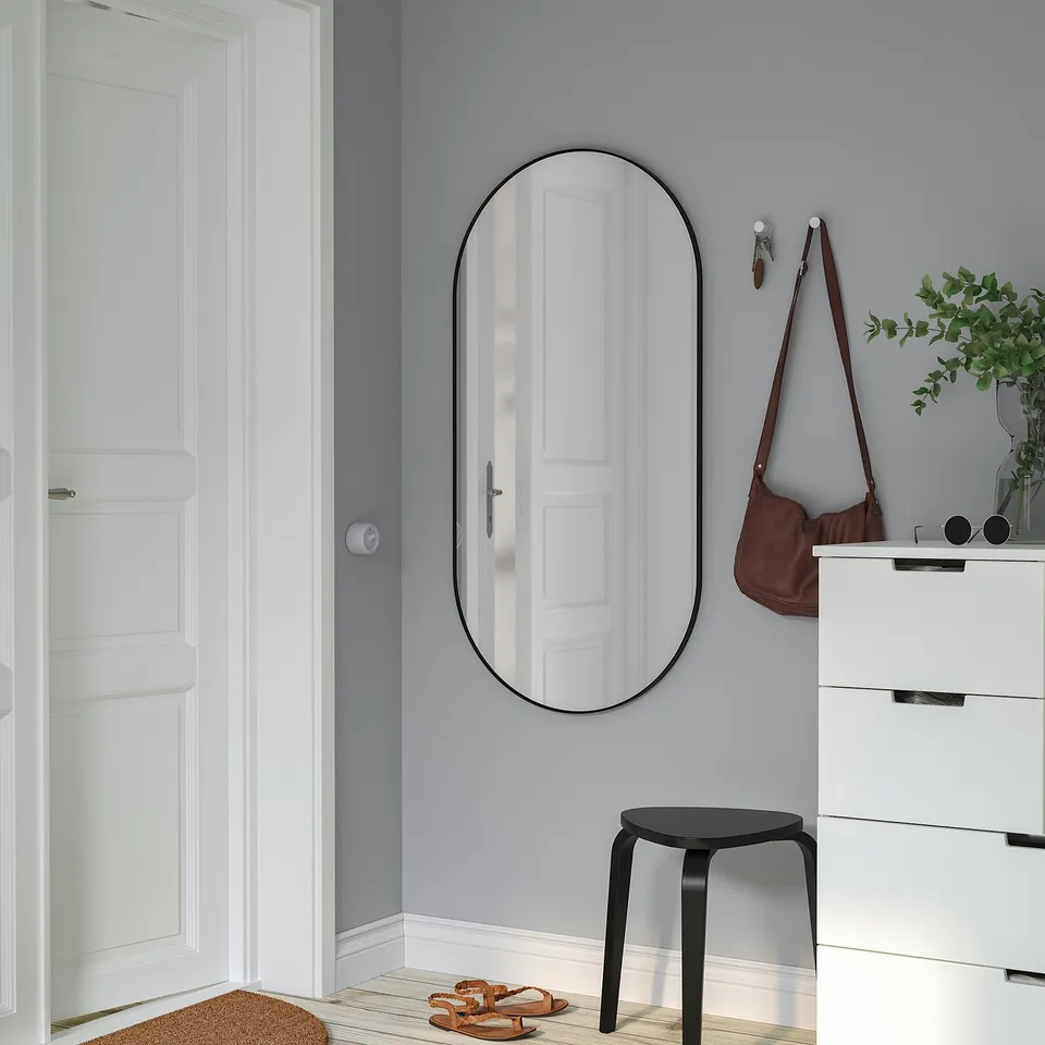 Minimalist entryway: large wall mirror, black frame, beside white dresser. Brown bag, sandals, small plant.