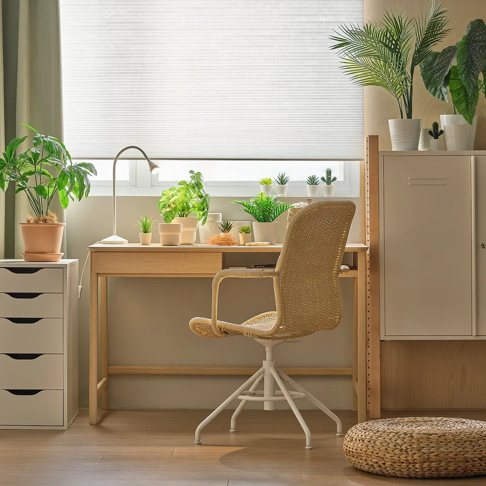 Home office desk with wicker chair. Desk topped with plants, light, and drawers. Chair adjustable, placed centrally. Blinds on window behind.