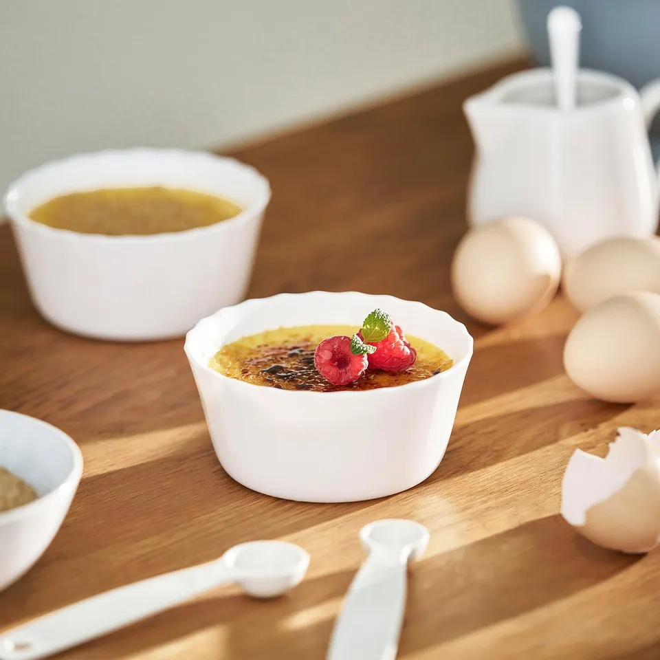 Ceramic ramekins with custard and berries on table, milk, eggs, and spoons.