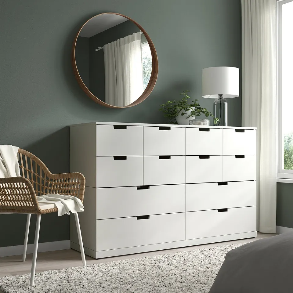 A modern bedroom with a white STOCKHOLM dresser, large walnut mirror, wicker chair, lamp, and plant. Dark green wall, sheer curtains.