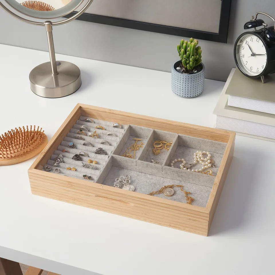 Wooden jewellery box with compartments holds rings, necklaces, and bracelets. Grey felt lining protects items. On a white desk near a brush and clock.