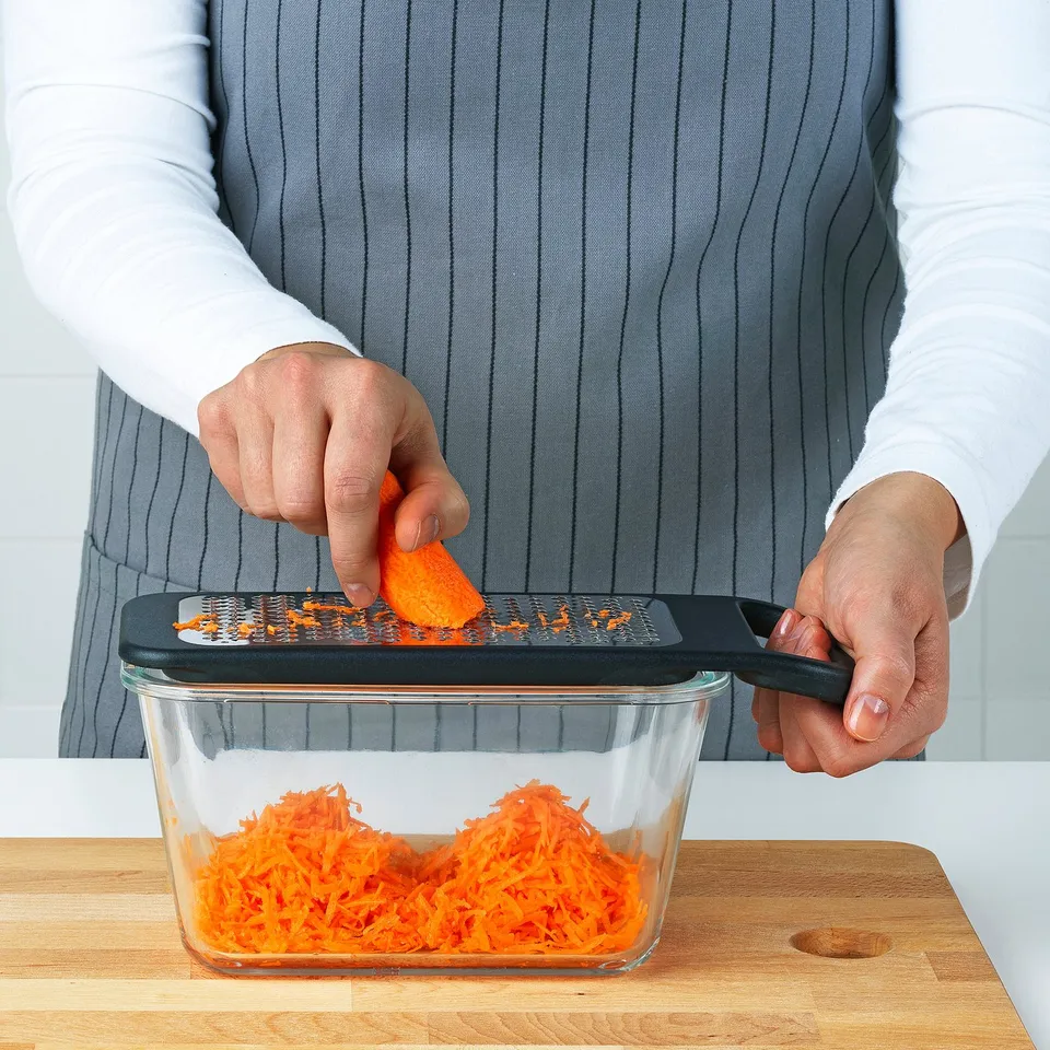 A person grates carrots using IKEA 365+ grater over a glass container, demonstrating its stability on edges.