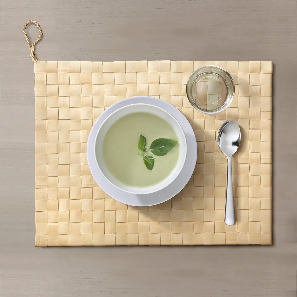A woven place mat displays a bowl of green soup and a spoon. The natural texture protects tables and reduces noise.