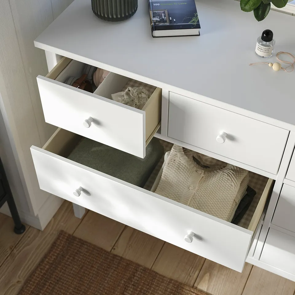 White dresser with three open drawers revealing neatly folded clothes. Top has a book, plant, and other items. Tapered legs.