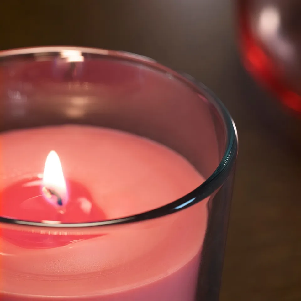 Close-up of a red scented candle in a glass holder, lit, revealing a white wick and smooth wax surface.