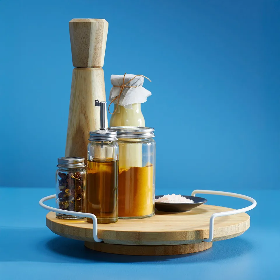 A round bamboo lazy susan with white handles holds jars of oils, spices, and a salt dish. A tall wooden pepper grinder stands beside it.