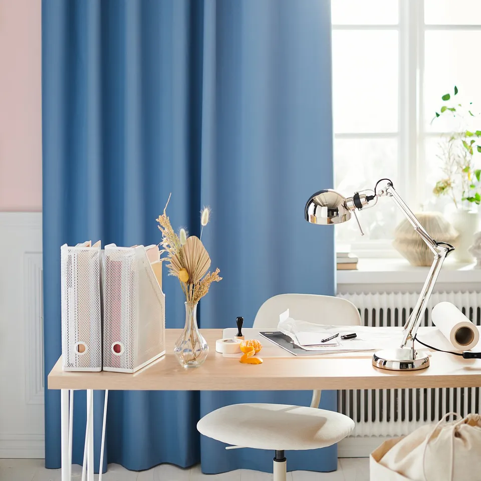 Modern office setup with blue curtains, wooden desk, white chair, metallic adjustable lamp, dried flowers in vase, and storage bins.