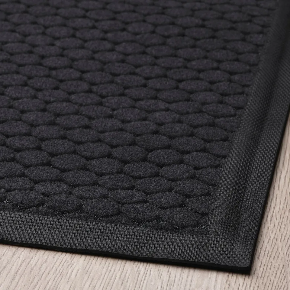 Black textured doormat on wooden floor, detailed diamond pattern, durable, recycled polyester, firm backing.