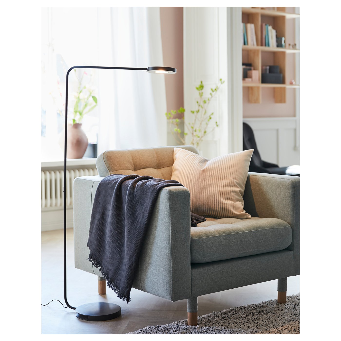 Cosy grey armchair with wooden legs, soft pillows, and a dark lamp beside it. Bright, modern living room setting with bookshelves and windows.