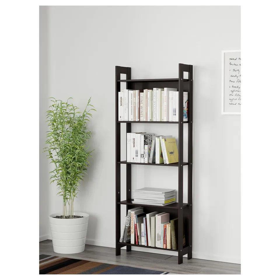 Black wooden bookshelf against a white wall, holding various books and magazines on four shelves. Beside it, a large potted green plant.