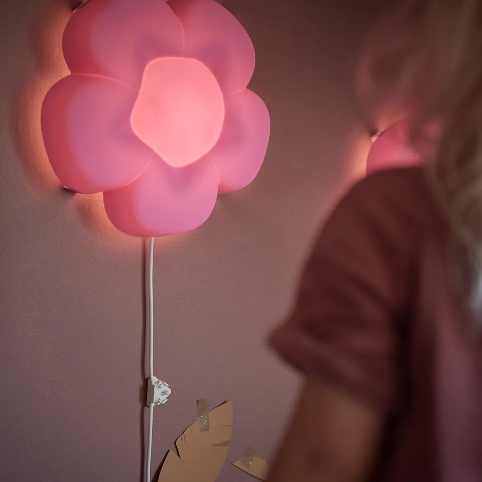 Pink flower-shaped wall lamp, made of 50% recycled plastic, softly lit, mounted on a wall.