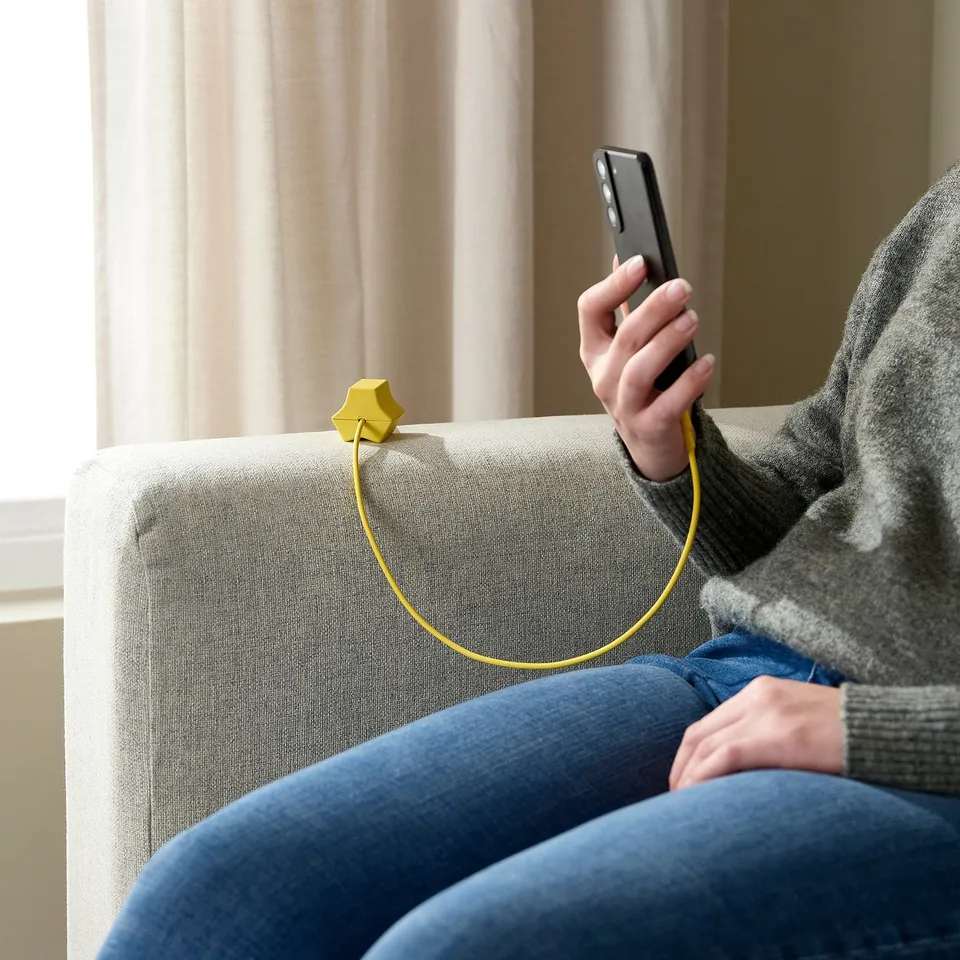 Person sitting on a couch, using a phone with a yellow HAVSKÅL usb anchor keeping the charging cable organised.