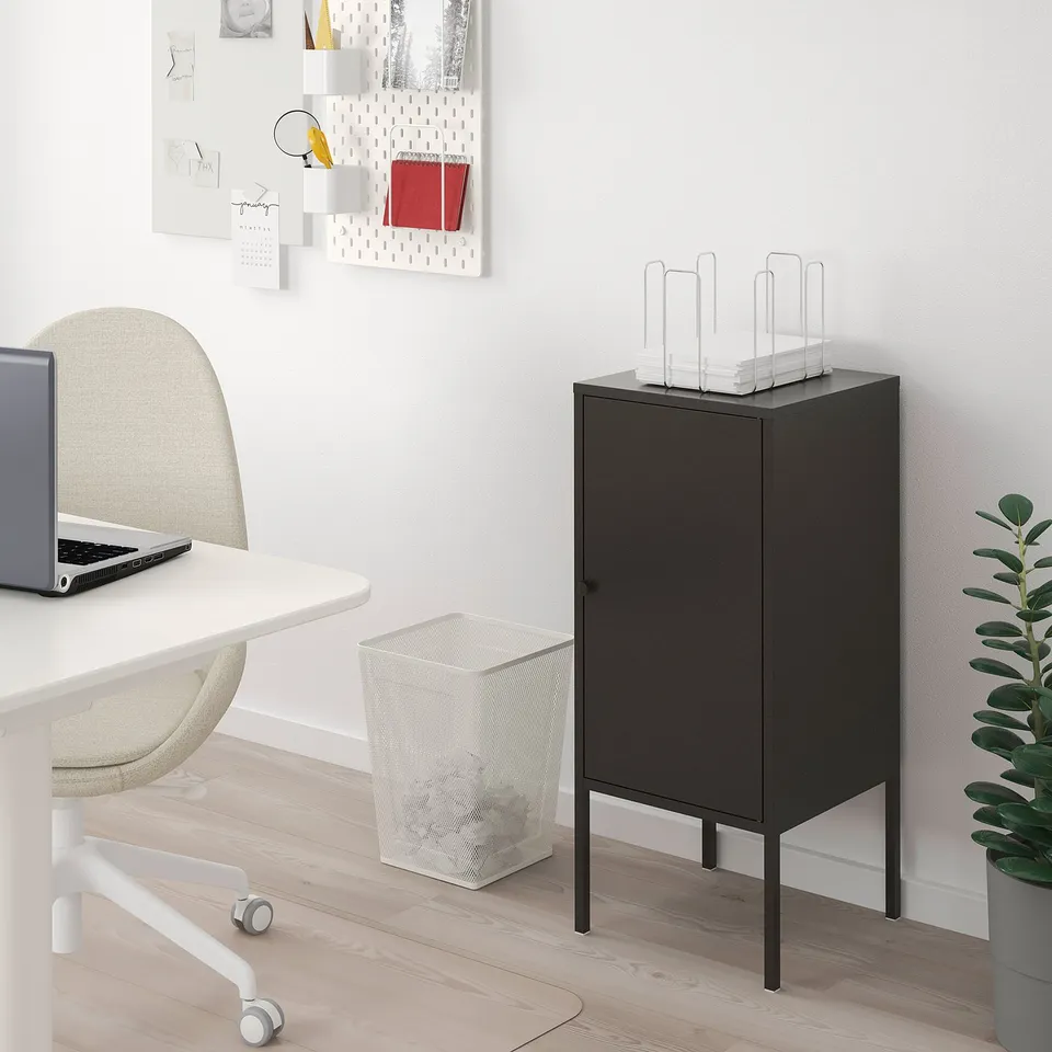 Black LIXHULT cabinet on thin legs beside desk, holding papers, smaller than desk and chair. White mesh bin, laptop, chair, and plant nearby.