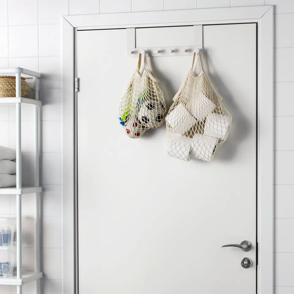 Two KUNGSFORS cotton bags hang on door, holding colourful items. White door and tiled wall. Nearby, shelving, towels, and wicker basket.