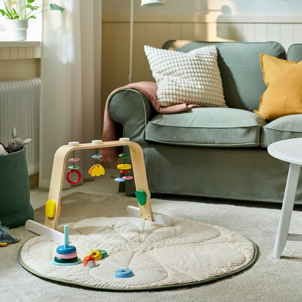 Living room with LEKA baby gym, light-colored arch with hanging toys, on a textured mat, near a green couch with colourful pillows.