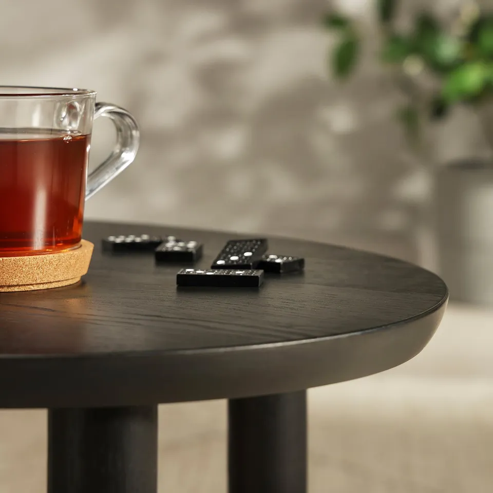 Glass mug with dark tea on a coaster beside three black remote controls on a dark round wooden table with a slightly shiny finish.