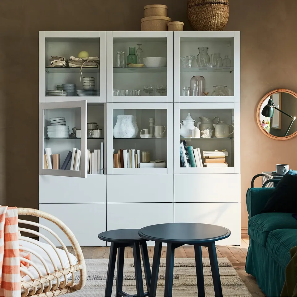 White SINDVIK cabinet with clear glass doors showcasing dishes, books, and vases. Accompanied by black tables and wicker chair.