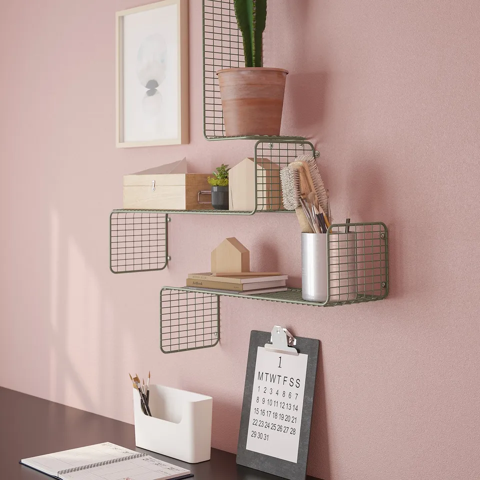Metal grid wall shelves display items creatively on pink wall. Shelves hold plant, brushes, books, and cup. Clipboard with calendar below.