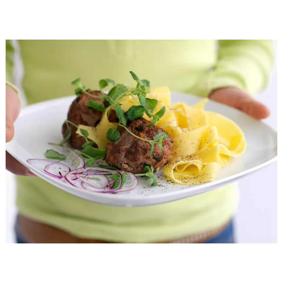 A person holding a square white porcelain plate with cooked meat, yellow sauce, noodles, and red onions garnished with green herbs.