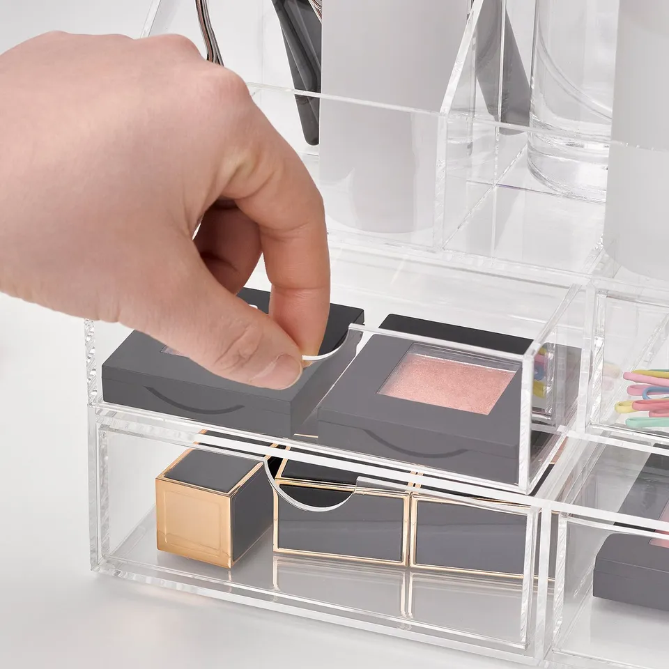 A hand reaching into a clear acrylic organiser with multiple compartments, holding makeup, including lipsticks and eyeshadow palettes.