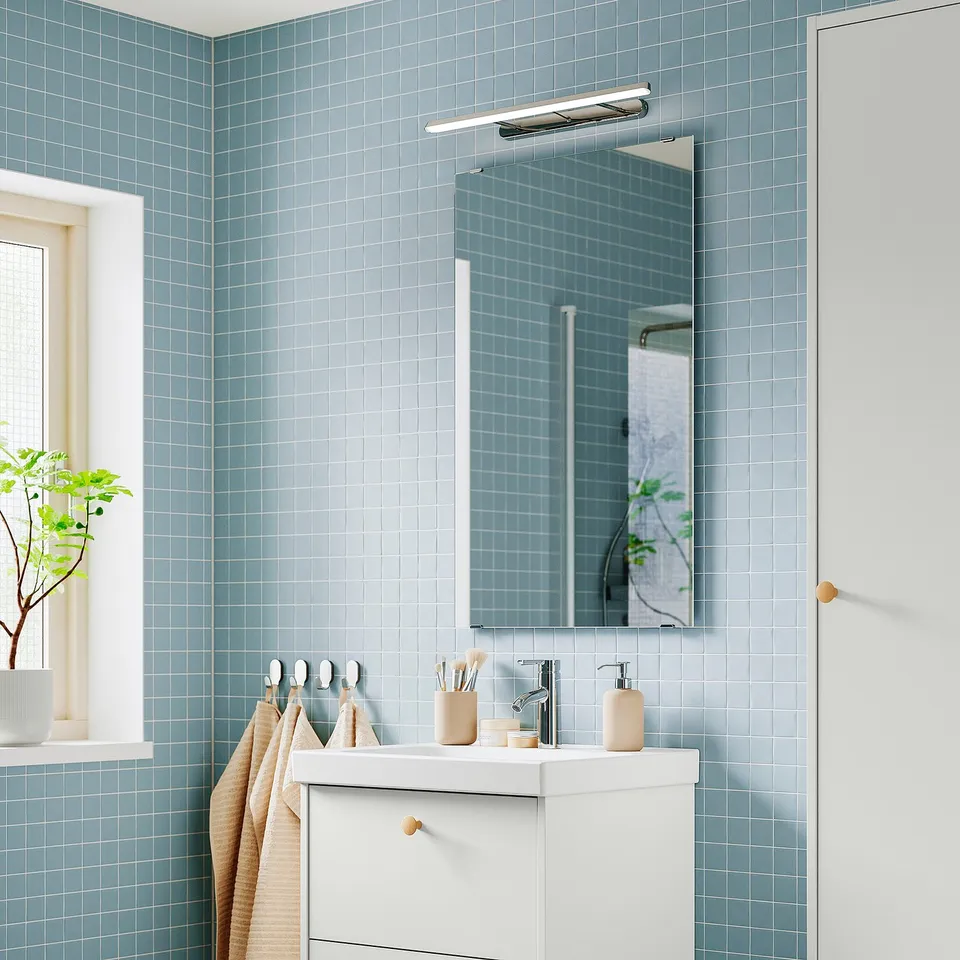 Modern bathroom with blue tiles, white vanity, mirror, FLASKPOST led light, small window, plant, and towels.