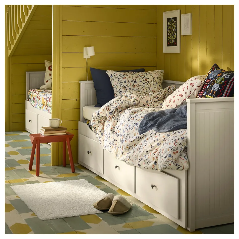 Bedroom with white storage beds, floral bedding, small red table, fluffy white rug, slippers, lamp, and wall art.