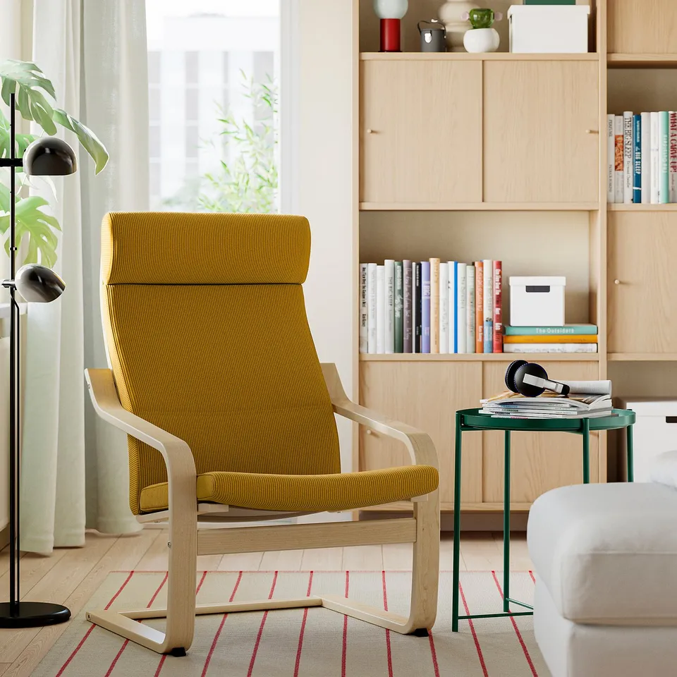 Modern living room with POÄNG mustard chair and cushion. Small table with headphones and magazines. Bookshelf with books and decor.