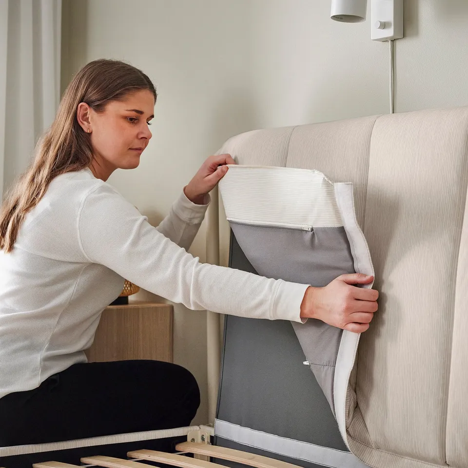 A person is fastening the zip on the removable, machine-washable headboard cover of a TÄLLÅSEN upholstered bed frame.