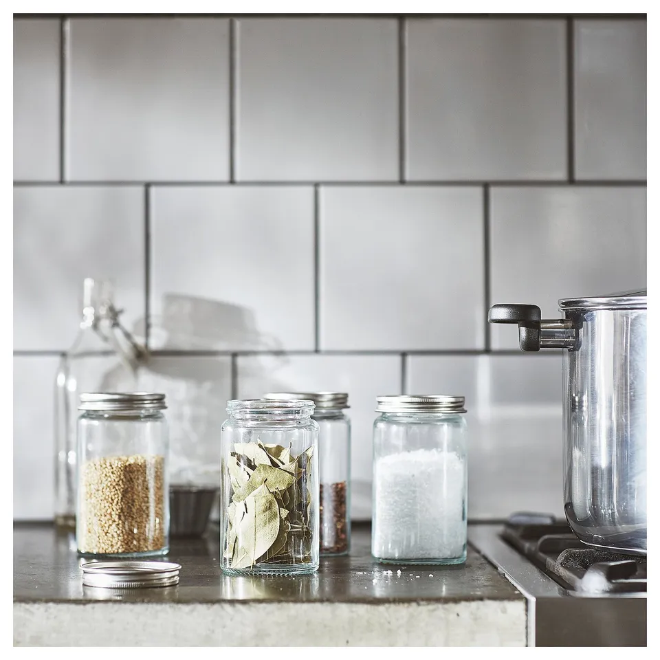 Glass spice jars with metal lids on counter, containing grains, leaves, salt. Minimalist kitchen design.