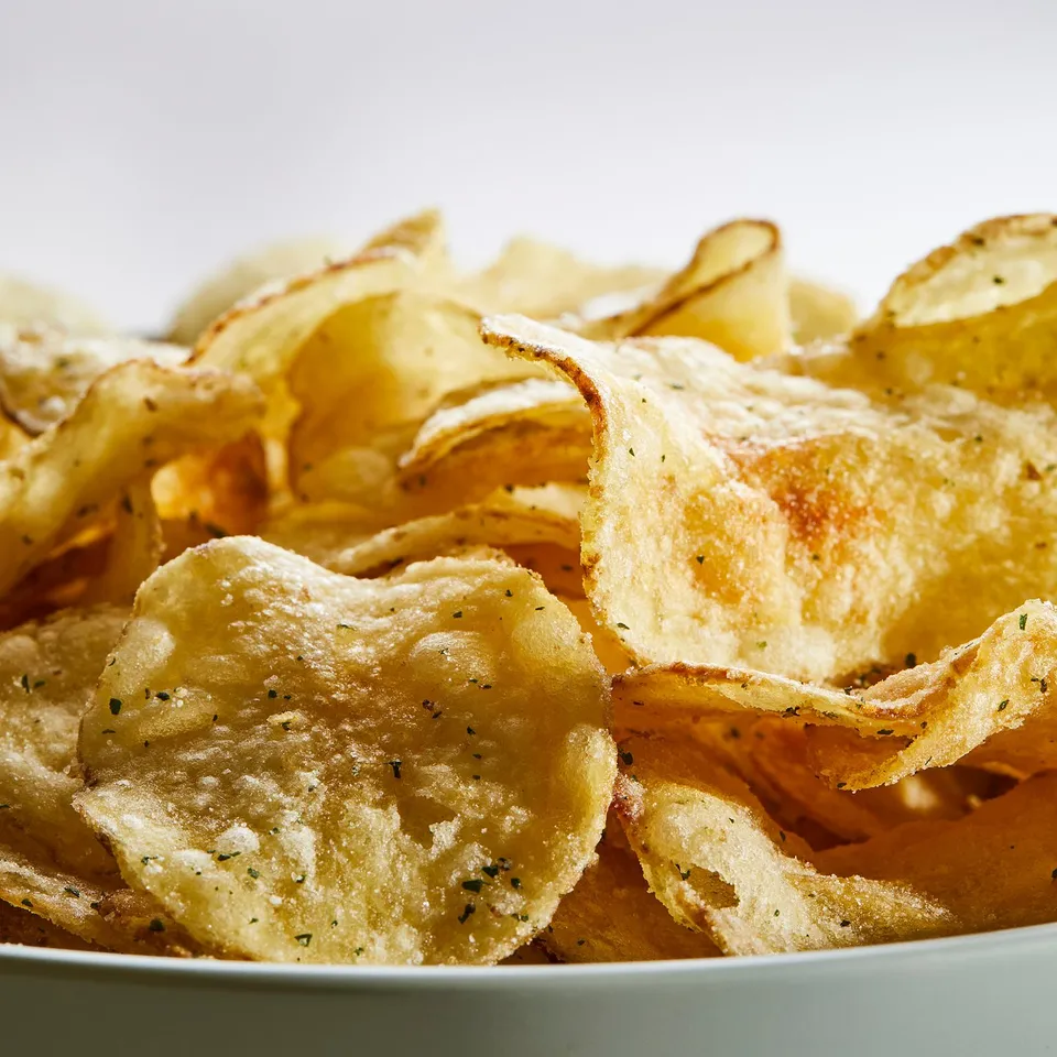 Pile of light golden potato crisps with visible seasoning, likely FESTLIGT brand, on a white surface.