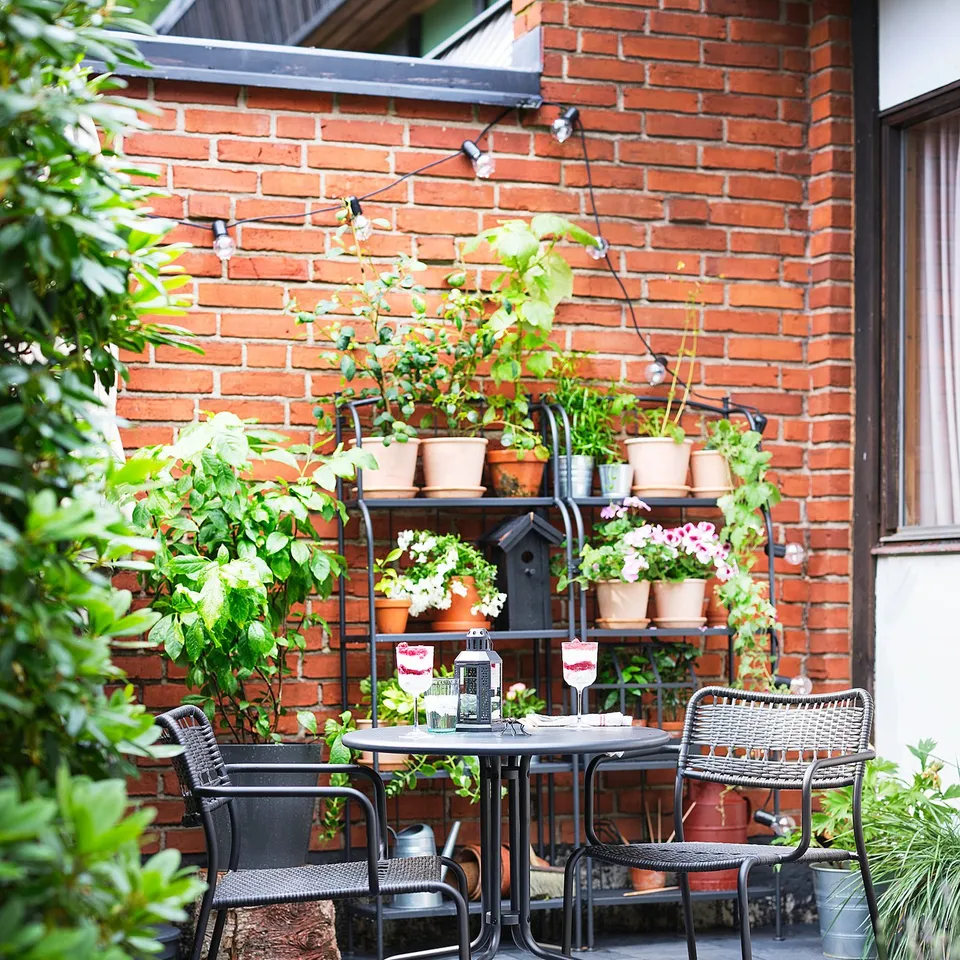 Cosy patio with LÄCKÖ black wrought-iron furniture against red brick wall, surrounded by lush green plants.