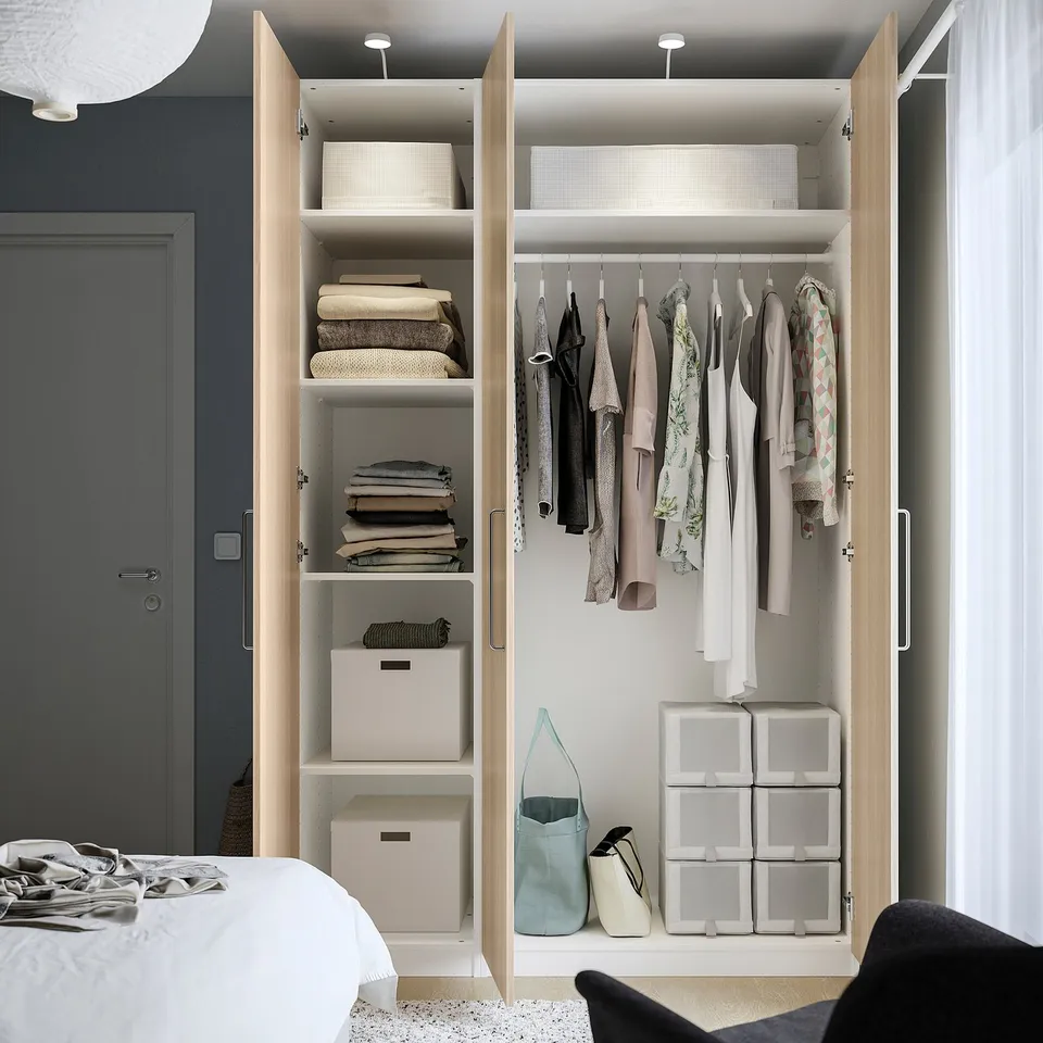 Open wooden wardrobe with shelves, folded clothes, hanging garments, storage boxes, and a blue tote bag.