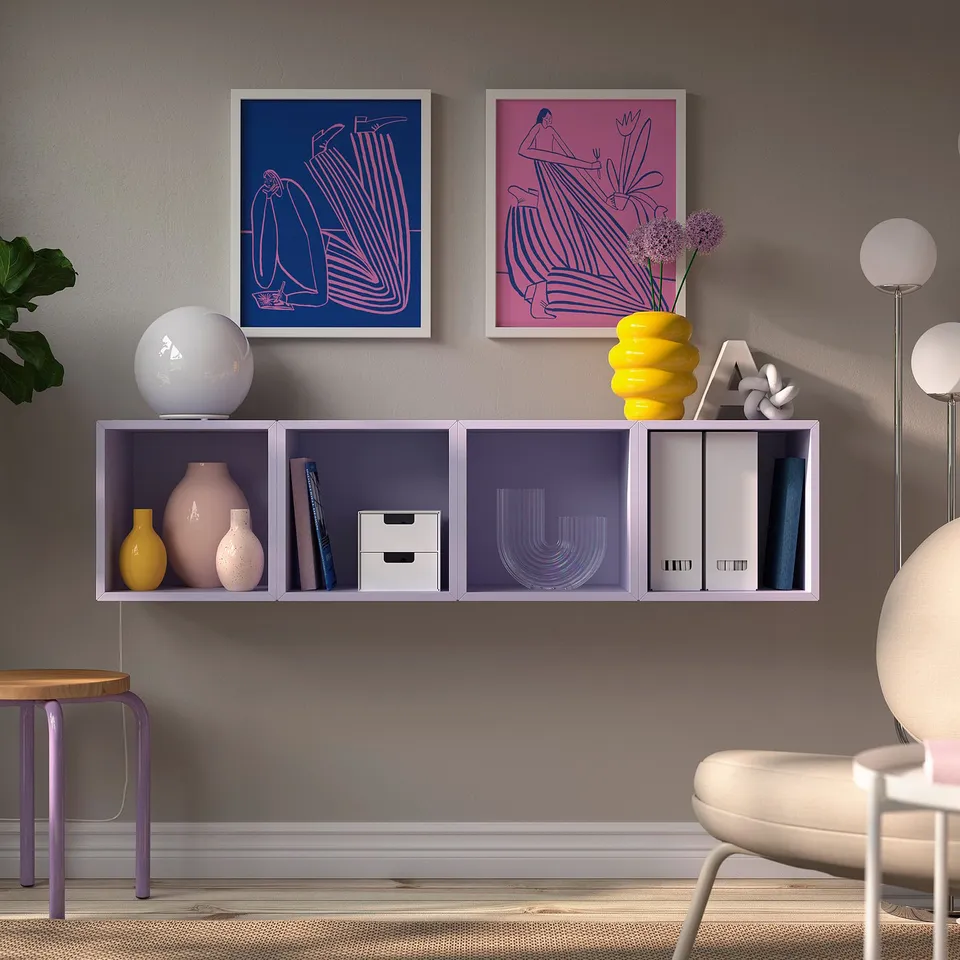 Small purple EKET wall unit, holding vases and books, between modern art prints, below a yellow vase and above a round chair.