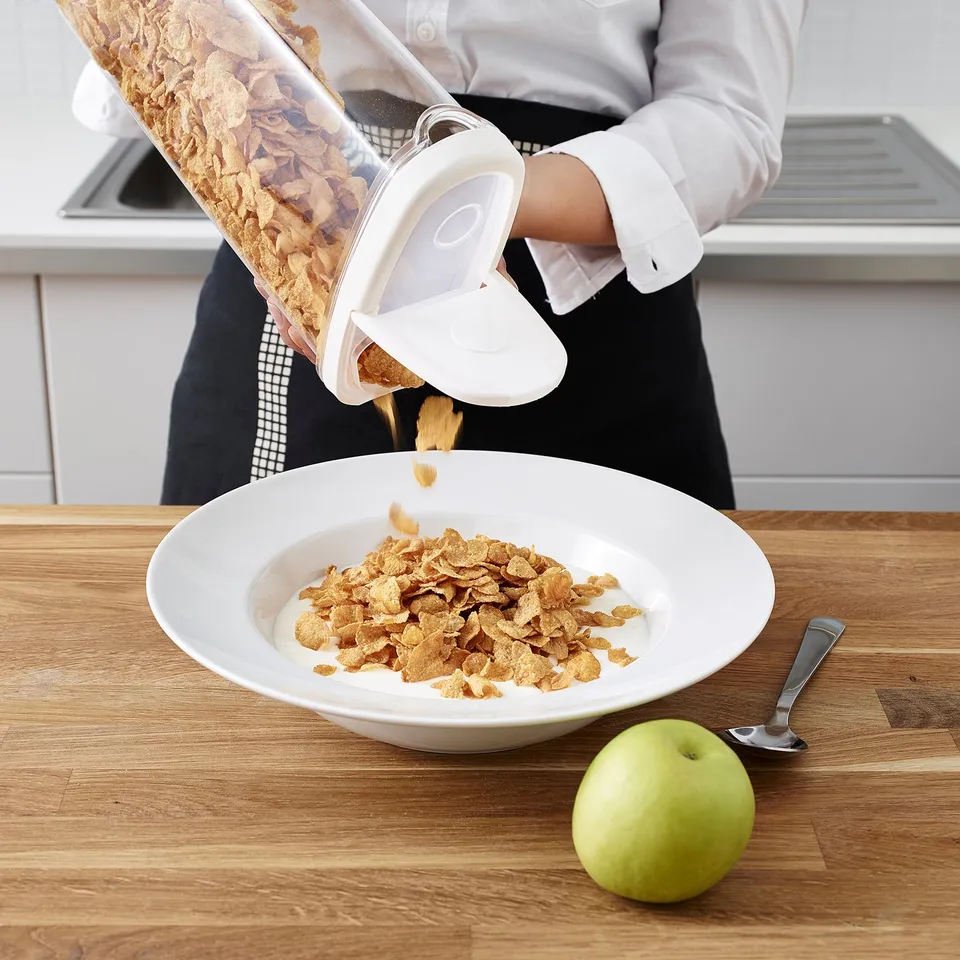 Person pouring cereal from a clear container into a white bowl. An apple and spoon sit beside the bowl on a wooden countertop.