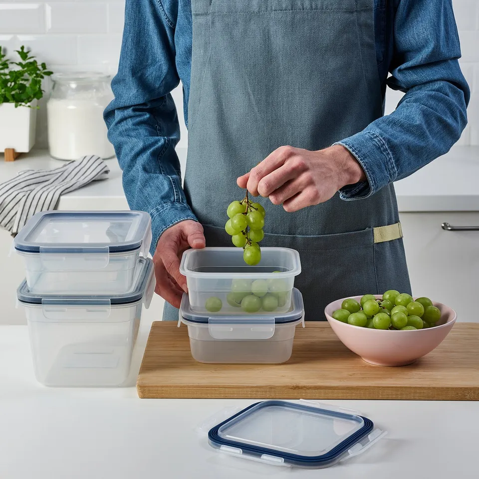 Person placing grapes in clear plastic food container on wooden board. Four containers with blue lids are stacked, one holds grapes.