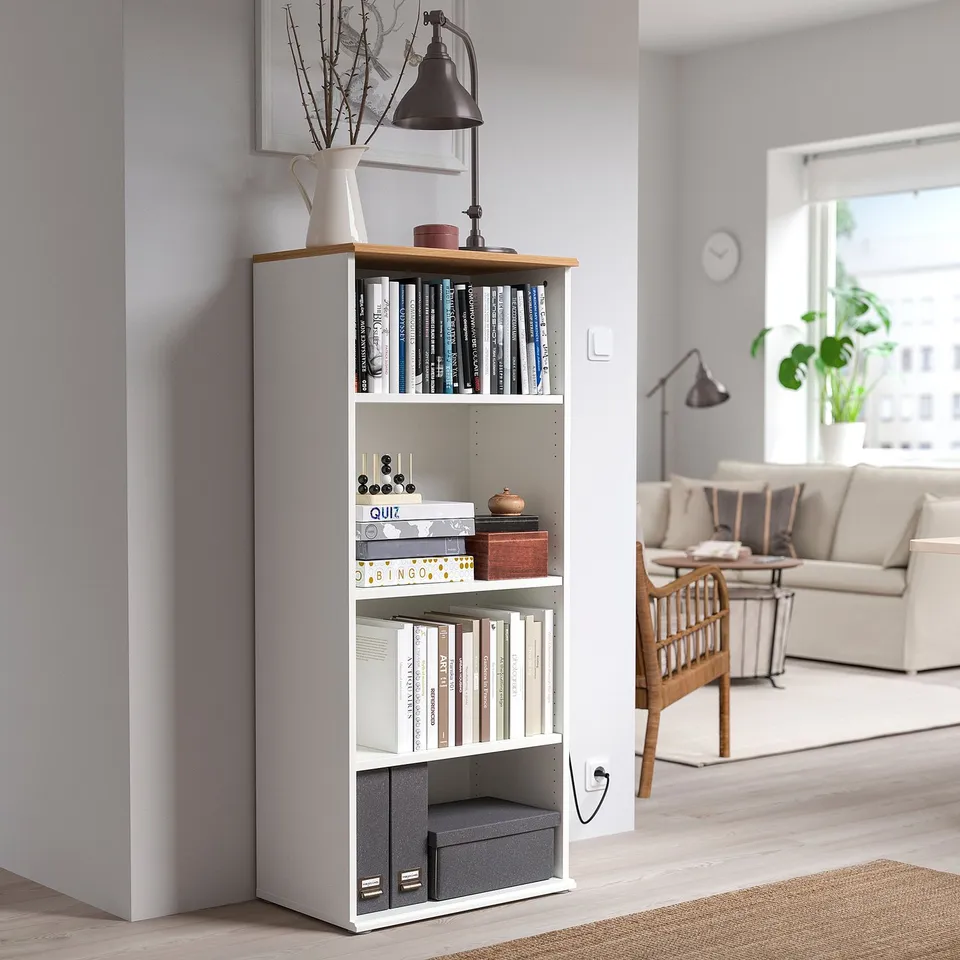 SKRUVBY white tall bookcase with a wooden top, storing books and decor in a living room, next to a window and a beige sofa.