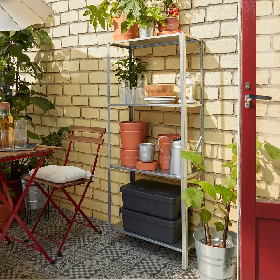 Balcony with HYLLIS silver shelving units storing plants and garden tools against cream brick wall.