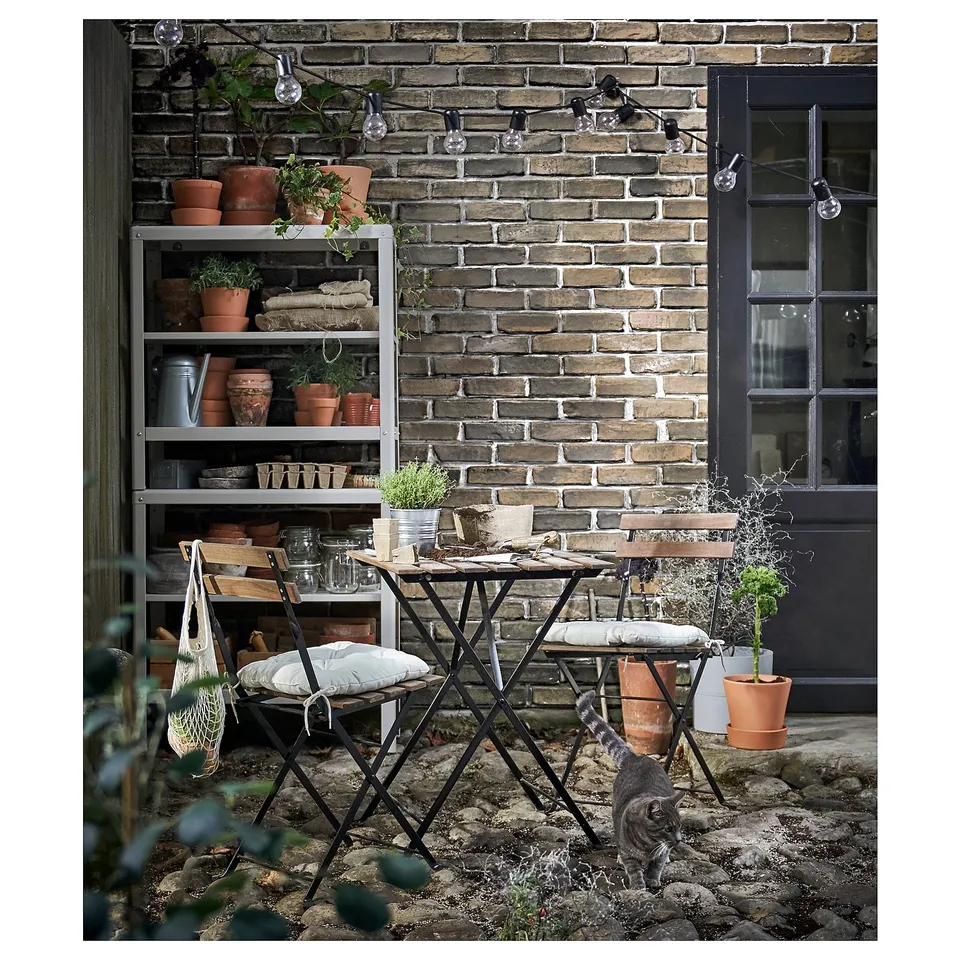Chrome KOLBJÖRN steel outdoor shelving unit with potted plants against a brick wall.