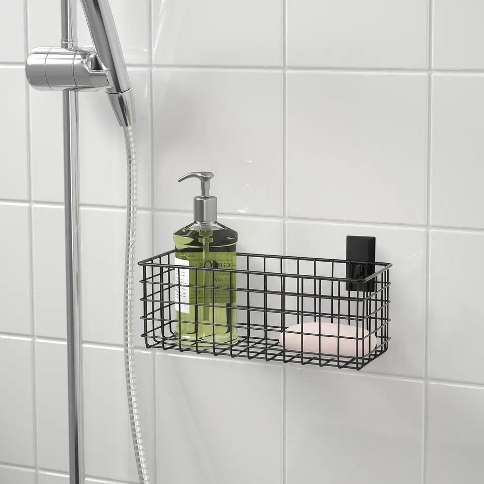 Black wire shower caddy holds green liquid soap dispenser next to shower head on white tiled wall.