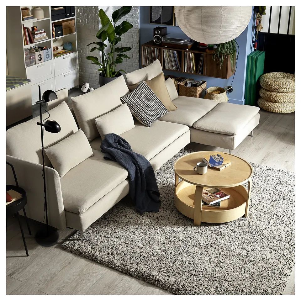 Modern living room: cream l-shaped sofa bed, wooden coffee table, black lamp, grey rug, and white brick wall.